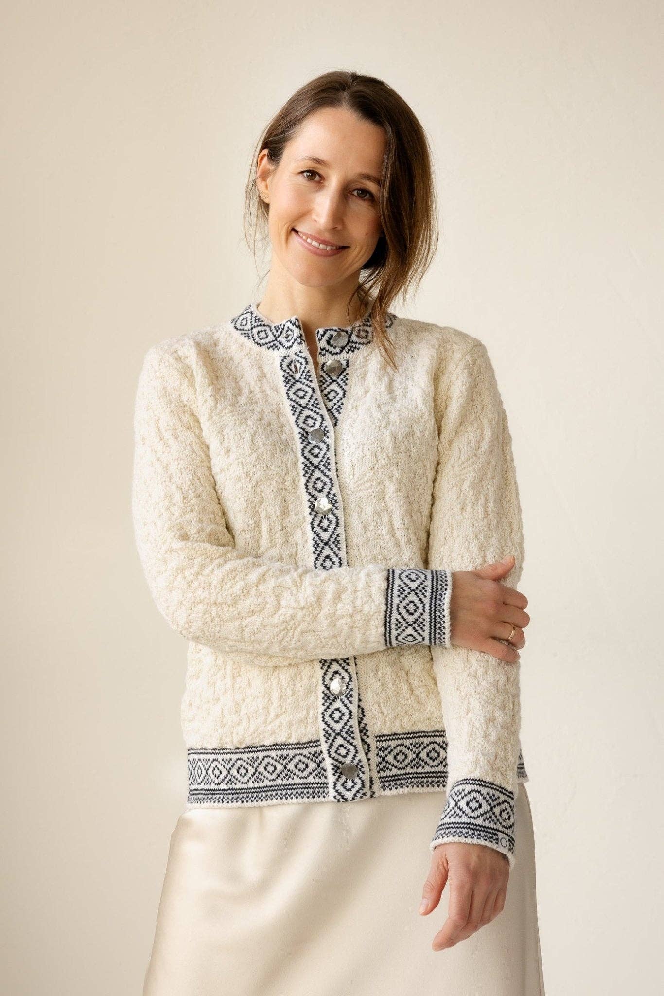 A woman stands smiling, wearing the Bryggen Women's Norwegian Cardigan (White) featuring black geometric patterns on the cuffs, collar, and edges, paired with a light-colored skirt.