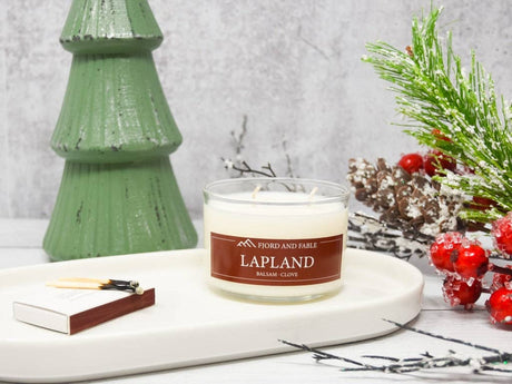 The Lapland Candle—Balsam Fir, Clove, & Cedar Blend (8 oz, 2 wicks) by Fjord & Fable sits on a white tray with matches, next to a green tree ornament and faux winter greenery decorated with red berries.