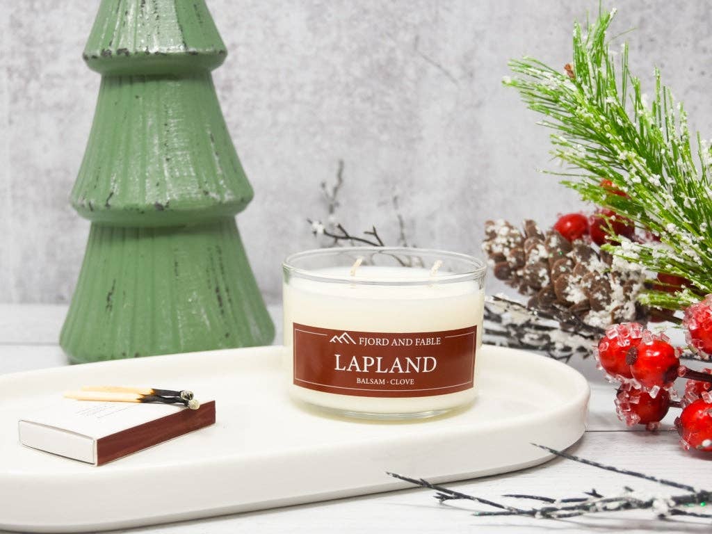 A decorative holiday scene includes a green tree ornament, the 4oz Lapland Premium Soy Candle with a Balsam Clove scent, a matchbox, and festive greenery adorned with red berries displayed on a white tray.