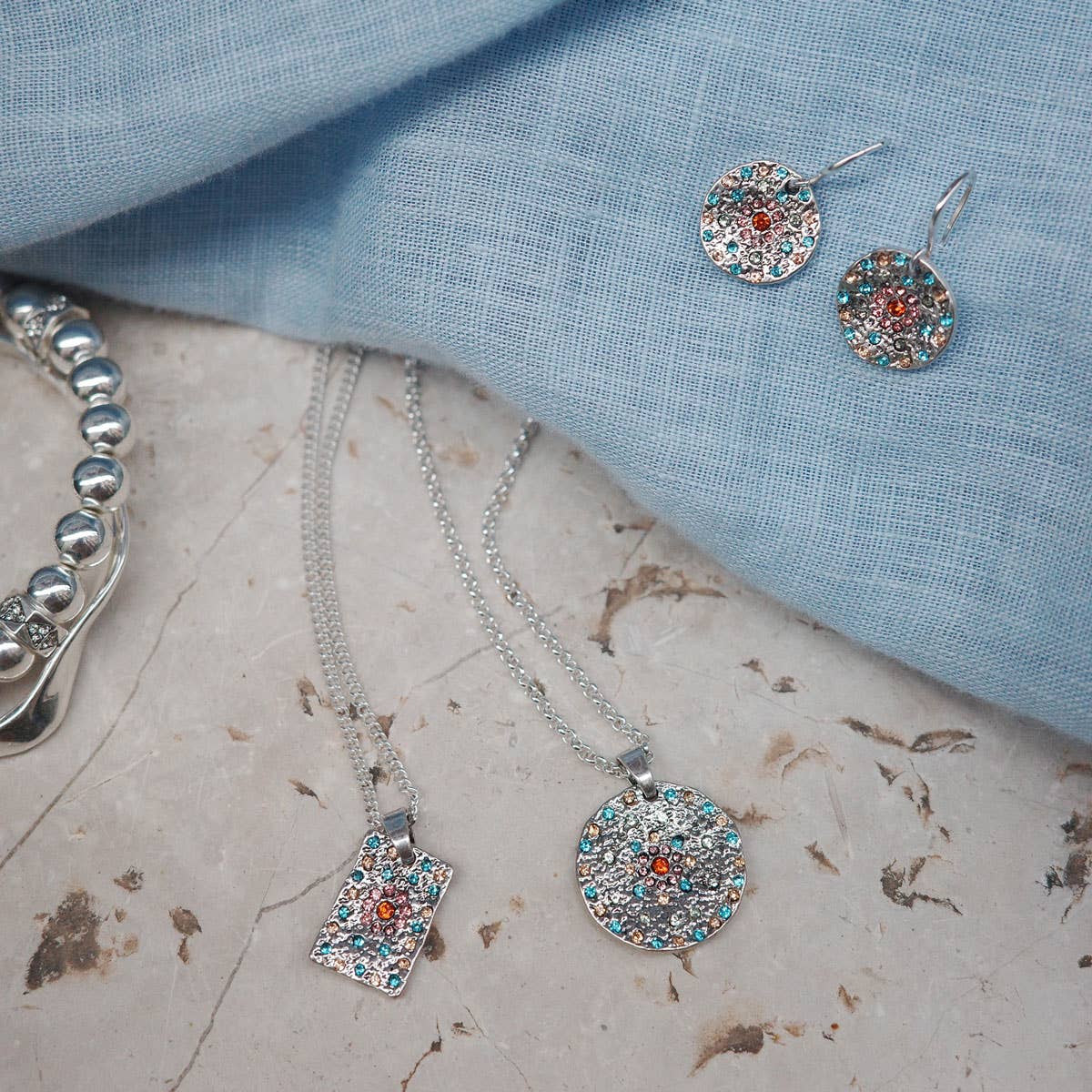 The A&C Oslo Vibrance Silver Pendant Necklace, adorned with colorful gemstones, is elegantly displayed alongside a matching bracelet and round earrings on marble and blue cloth.