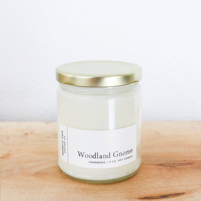 A Woodland Gnome Handmade Soy Candle made from natural soy wax comes in a glass jar with a gold lid, shown on wood with a white wall behind. Available as a 4oz tin or 8oz jar.