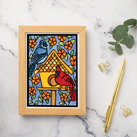 The Card: Birdhouse 5x7 Greeting Card by Sarah Angst features blue and red birds on a birdhouse amid orange flowers. Comes with an envelope and is blank inside. Made in America. Displayed on marble with a pen, binder clips, and green leaves.