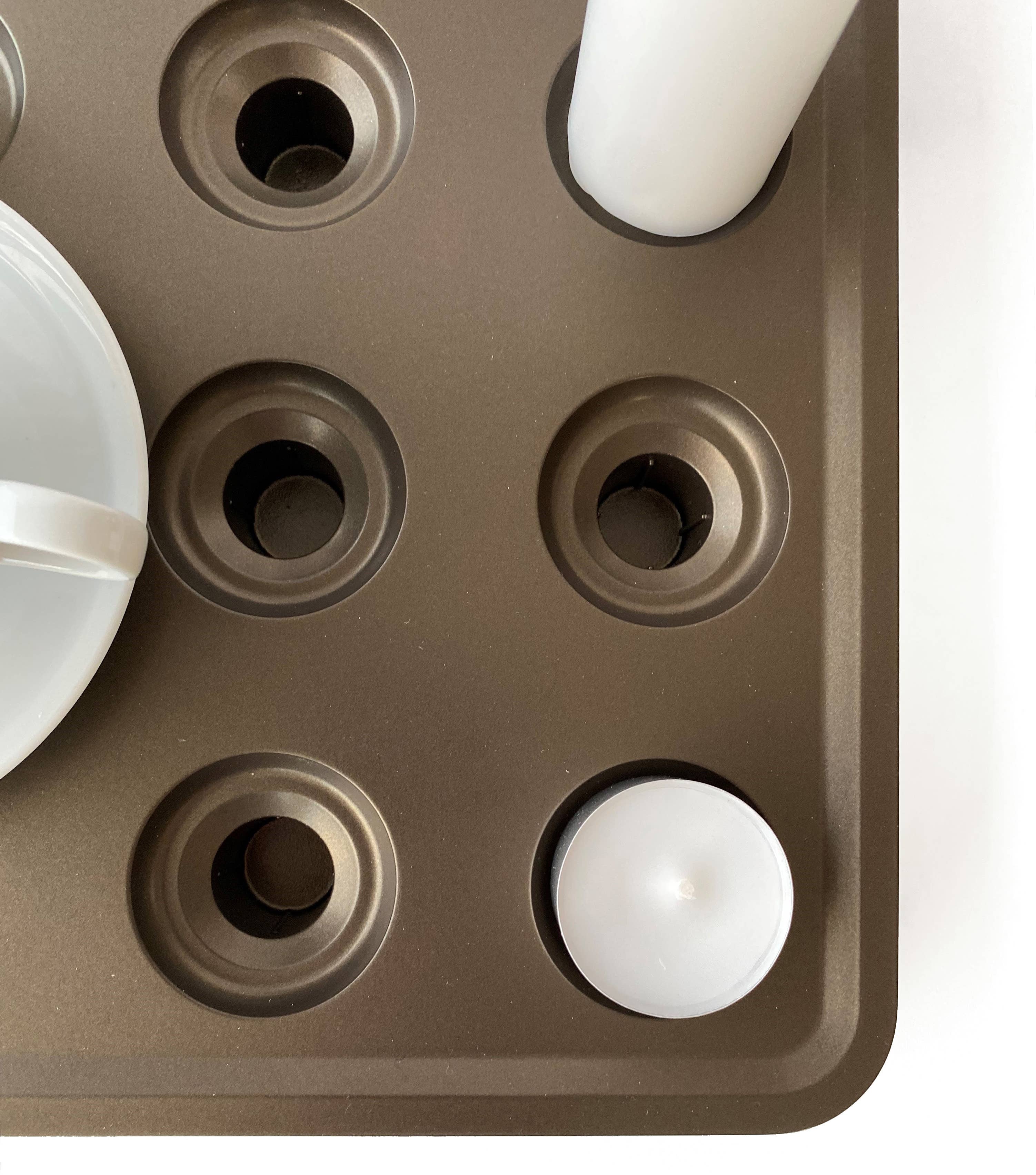 The Swedish-designed Stumpastaken White Powdercoated Candle Holder, crafted from recycled aluminum, is showcased as a brown muffin tray with nine slots, some holding white cylindrical candles, alongside a partly visible white cup with a handle.