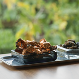 Chocolates in shiny copper foil and dark dried fruits on a birchwood tray, arranged on a wooden surface with Scandinavian design elements visible.