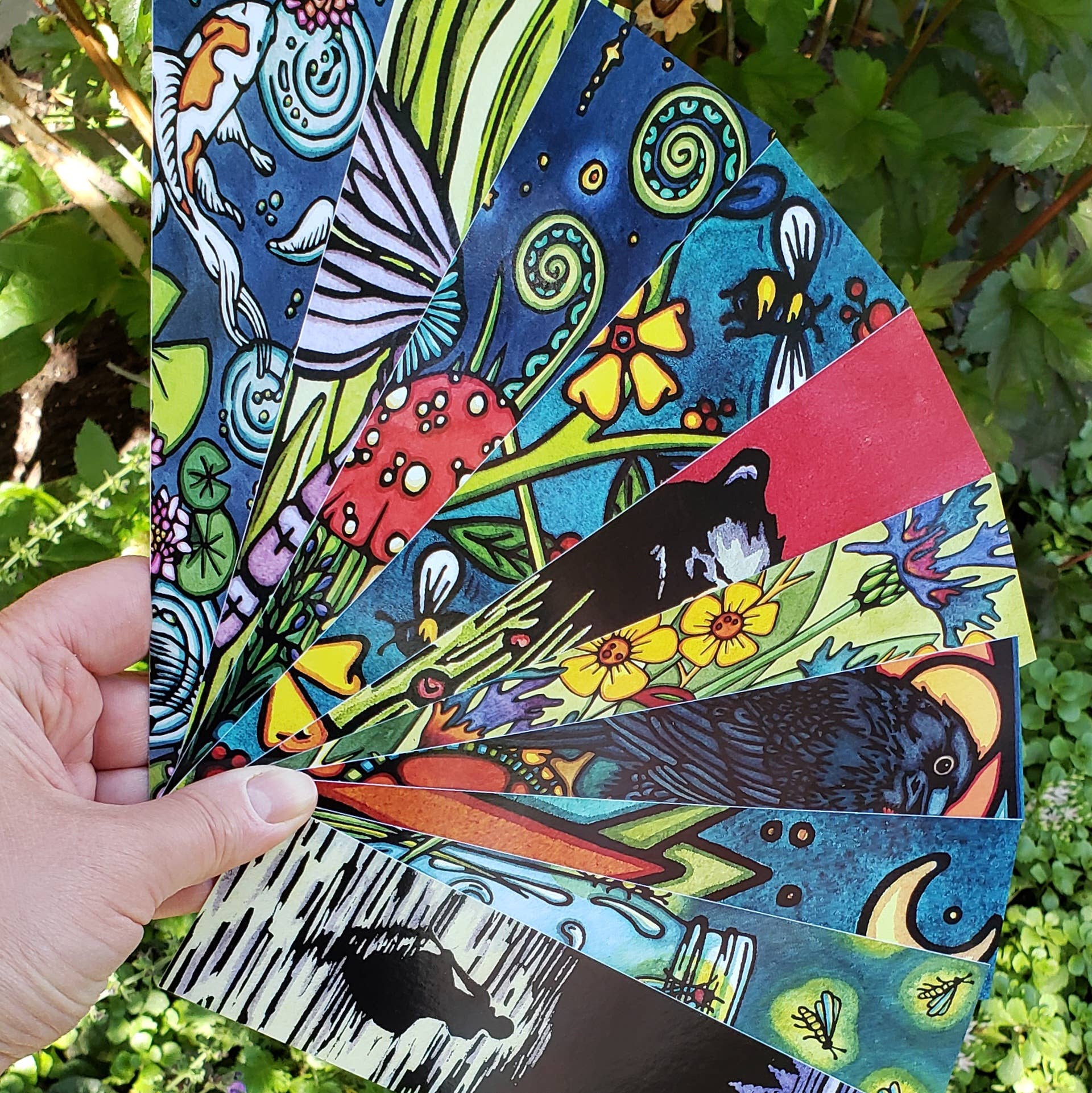 A hand holds a colorful fan adorned with nature-themed illustrations, reminiscent of the "Bookmark: Mushroom" by Sarah Angst, featuring elements like fish, flowers, birds, and insects. These vibrant details are captured on heavy cardstock with a matte gloss finish. In the backdrop lies tranquil greenery.