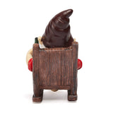 A garden gnome with a tall brown hat sits in a wooden chair, wearing a red shirt and holding a green bottle.