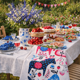 An outdoor table with cakes, strawberries, salads, and drinks is decorated with a floral centerpiece, blue napkins, festive cloth, bunting in the background, and a Tea Towel: Swedish Dala Horse Linen - Cotton Kitchen Towel by Lotta Spjute.
