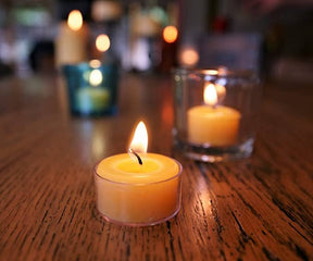 Handmade pure beeswax tea light candles from Candle: Pure Beeswax Tea Light Candle – Handmade Raw / Clear Cup glow warmly on a wooden table, their soft yellow flames flickering gently, creating a serene backdrop with blurred candles in the distance.