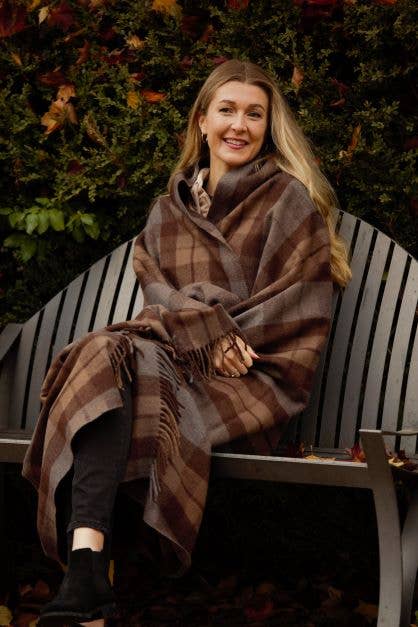 A woman sits on a bench outdoors, wrapped in a cozy Outlander Merino Wool Blanket, with autumn leaves and greenery in the background.