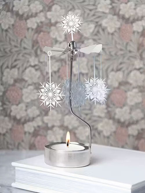 A delightful Pluto Design Angel Chime candle holder featuring elegant white snowflakes and stars embellishes a book. Its graceful design is perfectly festive against floral wallpaper, making it an ideal Christmas decoration.