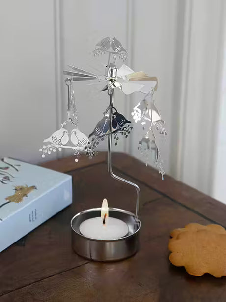 The Pluto Design Angel Chime Robin Rotary Candle Holder in silver—a small metal piece with robin-shaped ornaments—spins above a tealight on a wooden table beside a book and cookie, adding Scandinavian charm to your decor.