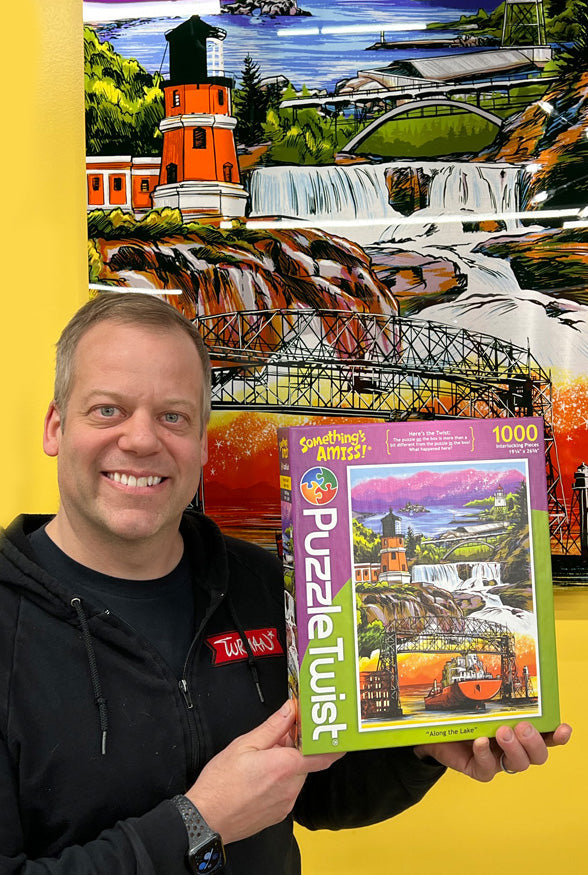 A person holding a PuzzleTwist: Along the Lake (1,000 Pieces) box featuring a lighthouse and waterfall image, perfectly matching the Adam Turman artwork in the background.