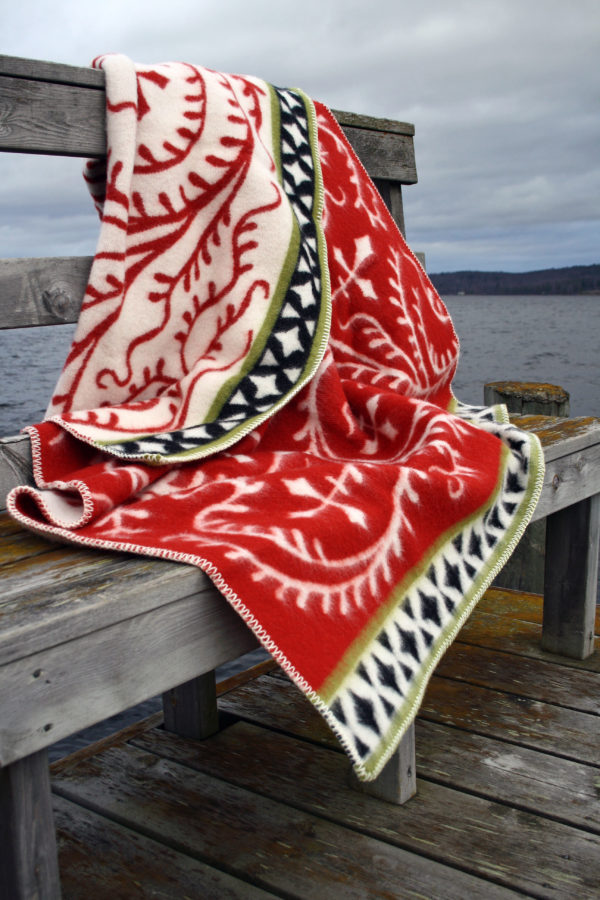 The Albertus Red wool blanket by Kerstin Landström, made of 100% lambswool, draped over a wooden bench on a dock beside calm water under a cloudy sky.