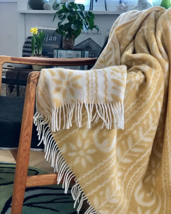 A wooden chair draped with the Anno 1862 blanket, made of 75% Norwegian lambs wool, 15% merino wool, and 10% silk by Kerstin Landstrom, sits in a room. Behind it are a potted plant and a small daffodil on a shelf.
