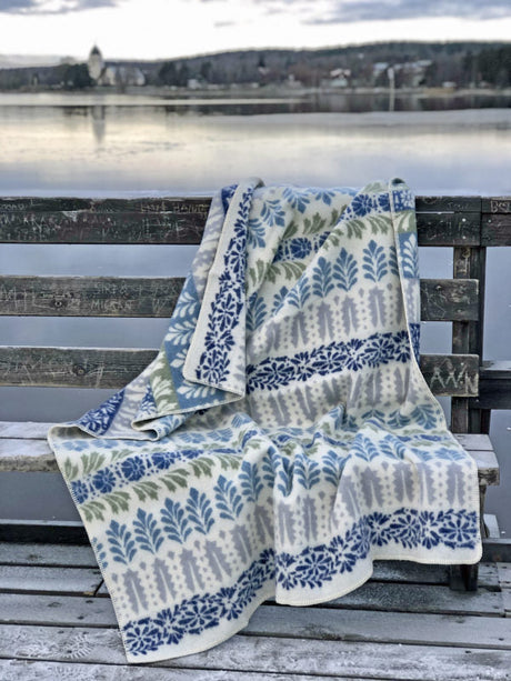 An Arvet Heritage blue patterned blanket, made from 100% Norwegian lambs wool by Kerstin Landstrom, draped over a weathered wooden bench by a lake under a cloudy sky.