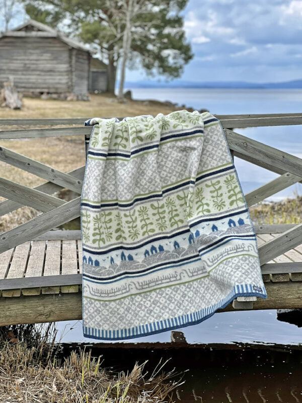 A Bohus Summer blanket by Kerstin Landstrom, made from 100% Norwegian lambswool with green, blue, and gray patterns, is draped over a wooden fence by the lake. A cozy cabin and tall trees stand in the background under a cloudy sky. OEKO-TEX certified for sustainable quality.