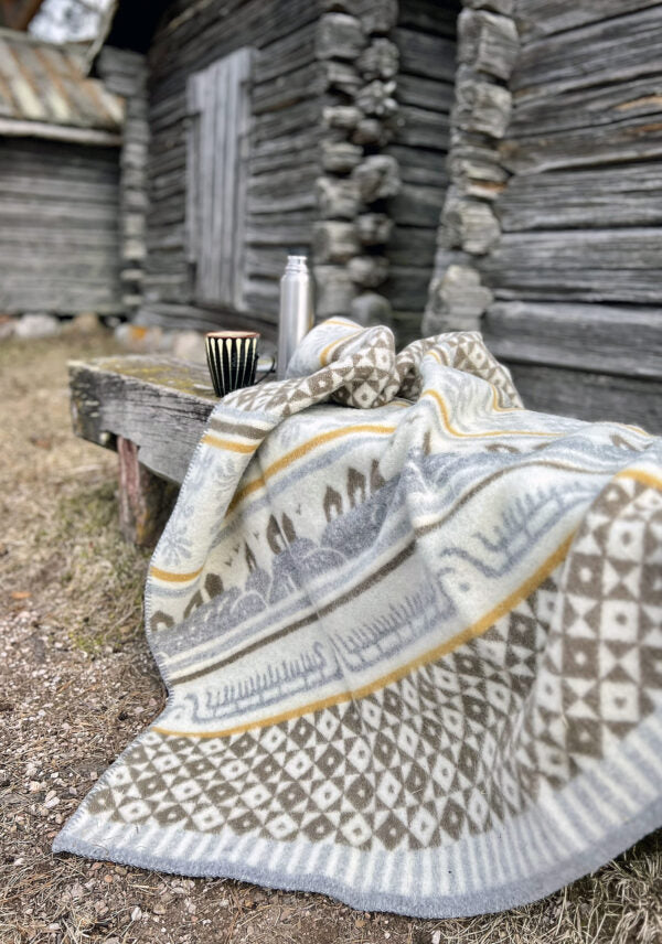 A Bohus blanket made of 100% Norwegian lambs wool by Kerstin Landstrom drapes over a wooden bench, paired with a thermos and cup. The serene scene is set against the rustic charm of an old wooden building.