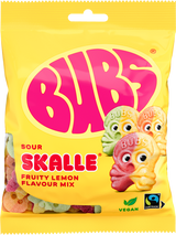 A yellow 90g bag of Faire - Sweet as Fudge Candy: Bubs Sour Foam Fruity Lemon Skulls shows three cartoon lemon skull gummies, vegan and Fairtrade logos, pink BUBS branding, and Scandinavian candy pieces visible through the packaging.