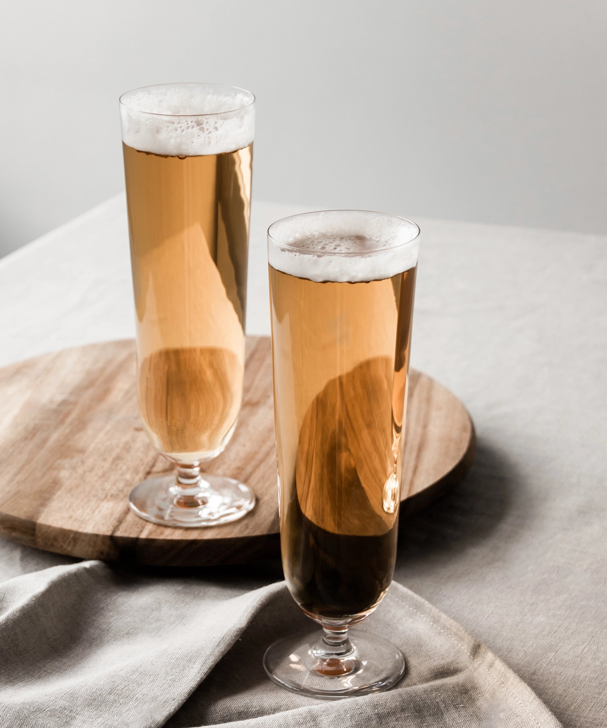 Two Orrefors Beer Pils 15.5oz glasses, designed by Erika Lagerbielke, hold amber beer as they rest elegantly on a wooden tray covered with a linen cloth.