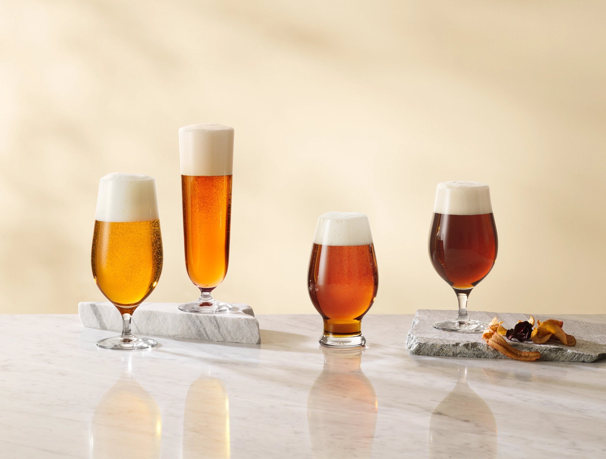 On a marble surface, four 15.5oz Orrefors Beer Pils glasses sit elegantly crafted by Erika Lagerbielke. Each frothy-topped glass is accented with dried leaves on the two right, perfect for savoring a classic Beer Pilsner experience.