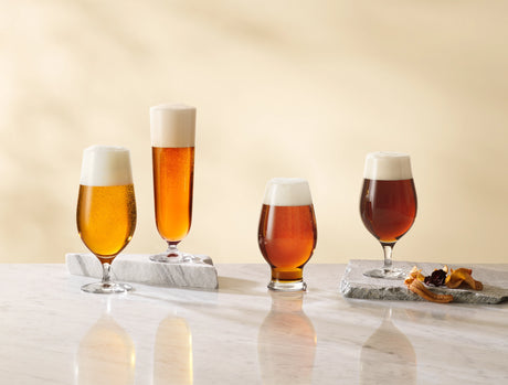 On a marble surface, four 15.5oz Orrefors Beer Pils glasses sit elegantly crafted by Erika Lagerbielke. Each frothy-topped glass is accented with dried leaves on the two right, perfect for savoring a classic Beer Pilsner experience.