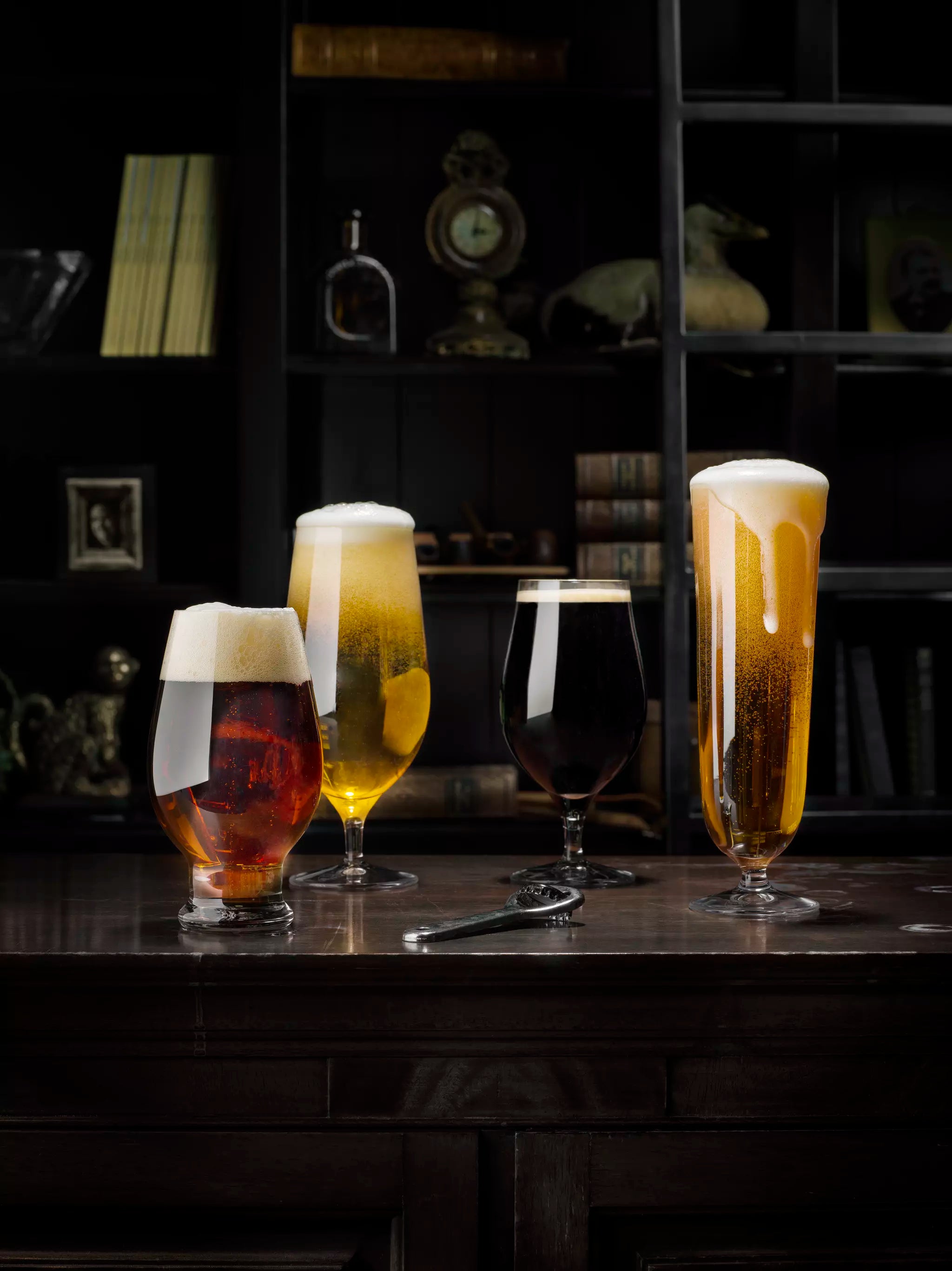 Four Orrefors: Beer Pils 15.5oz glasses sit on a wooden table alongside an Orrefors bottle opener. The backdrop features dark shelves filled with books and decorative items, creating a cozy ambiance.