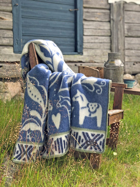 A Dalecarlia blanket, blue and white with Dala horse designs by Kerstin Landstrom, is made from 100% Norwegian Lambs Wool. Its draped over a wooden chair on grass, next to a rustic wall and metal milk can.