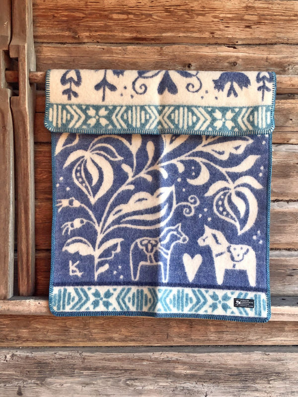 The DALARNA Baby Blanket—made from 100% organic Norwegian lambswool and featuring blue and white horse and floral motifs—hangs on a rustic wall, making a charming Swedish heritage gift for babies (65 × 95 cm).