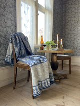 A wooden chair with a Kerstin Landstrom Blaklint Cornflower blanket of blue and white 100% Norwegian lambs wool rests next to a small round table with lit candles, potted plants, and glass bottles. The room showcases floral wallpaper and classic wooden flooring.
