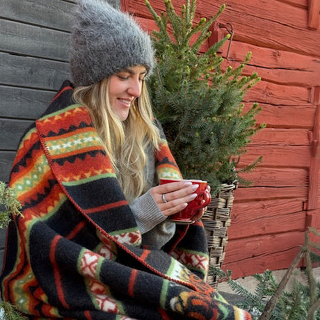 A woman in a gray knit hat sits outside, holding a red mug beside evergreen branches and a red wall, wrapped in the Bunad by Kerstin Landstrom Design Sweden 100% Norwegian Wool Blanket.