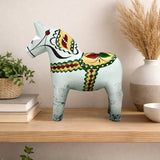 An Antique Inspired Swedish Dala Horse in white with gray feet (17cm) is displayed on a wooden shelf alongside books, a potted plant, and a vase of dried grass.