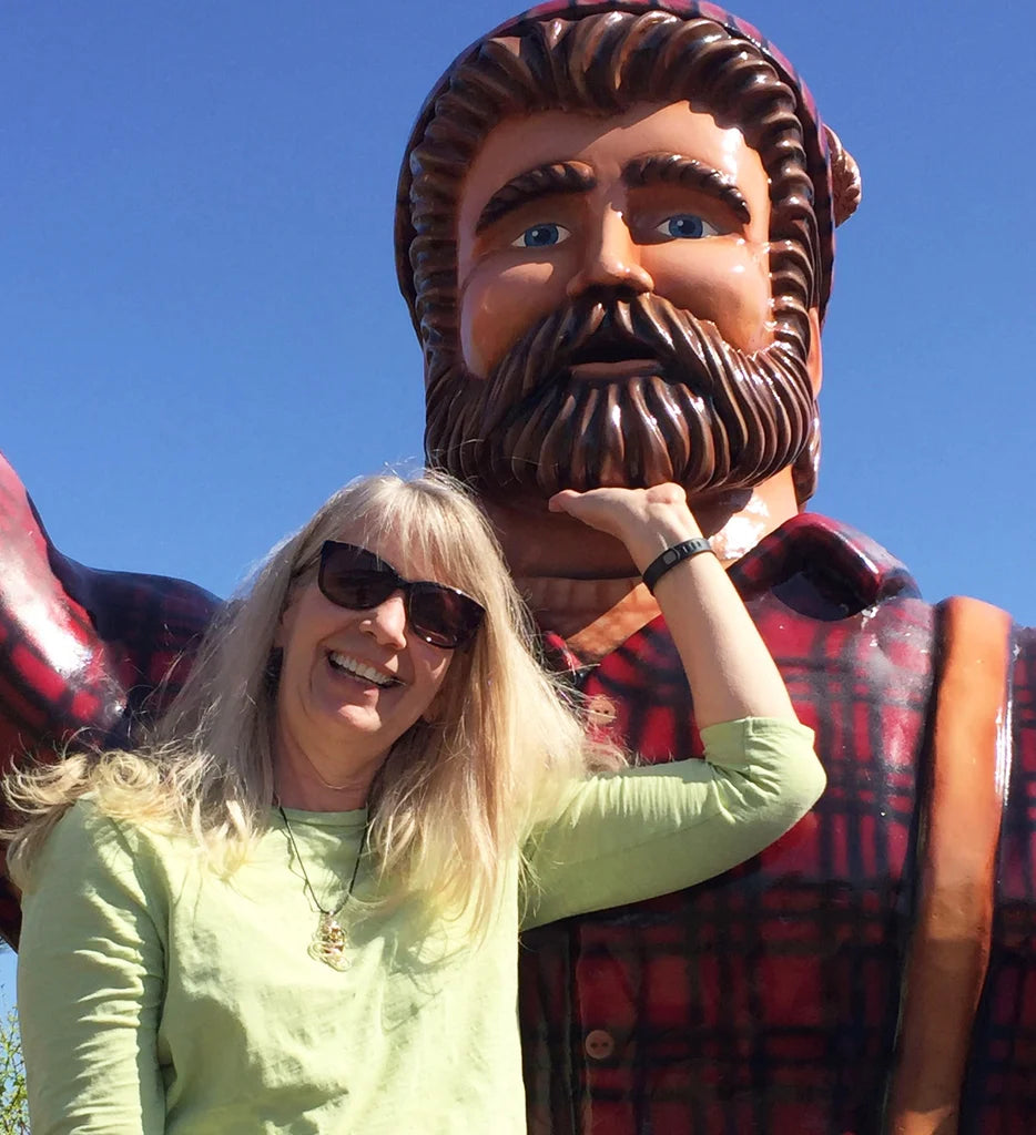 A woman in sunglasses stands next to a large Paul Bunyan statue dressed in a plaid shirt, with her hand resting on the statue’s chin.