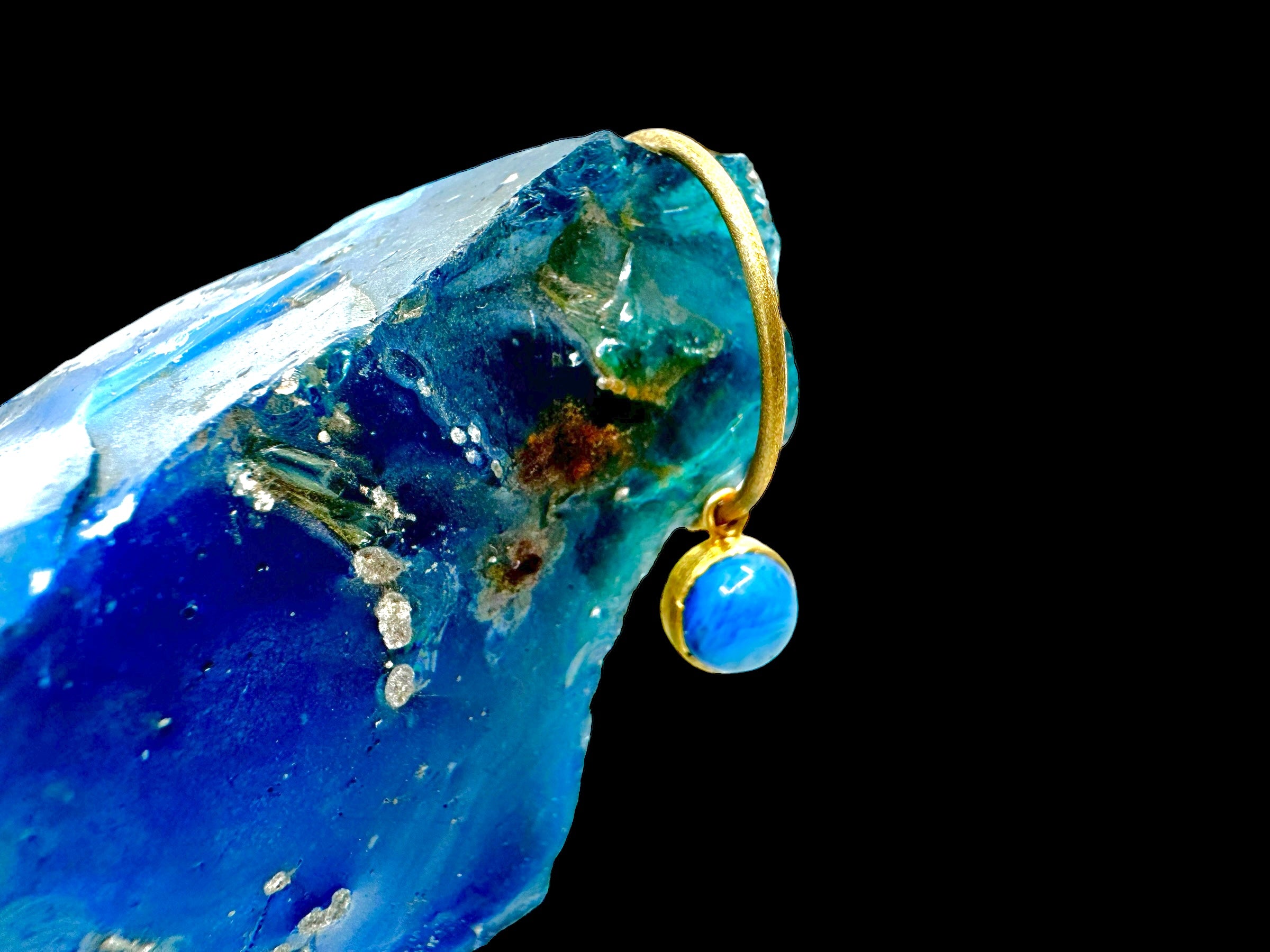 A pair of gold-plated earrings with Swedish Blue stones delicately hang against a rough, translucent blue gemstone on a black background.