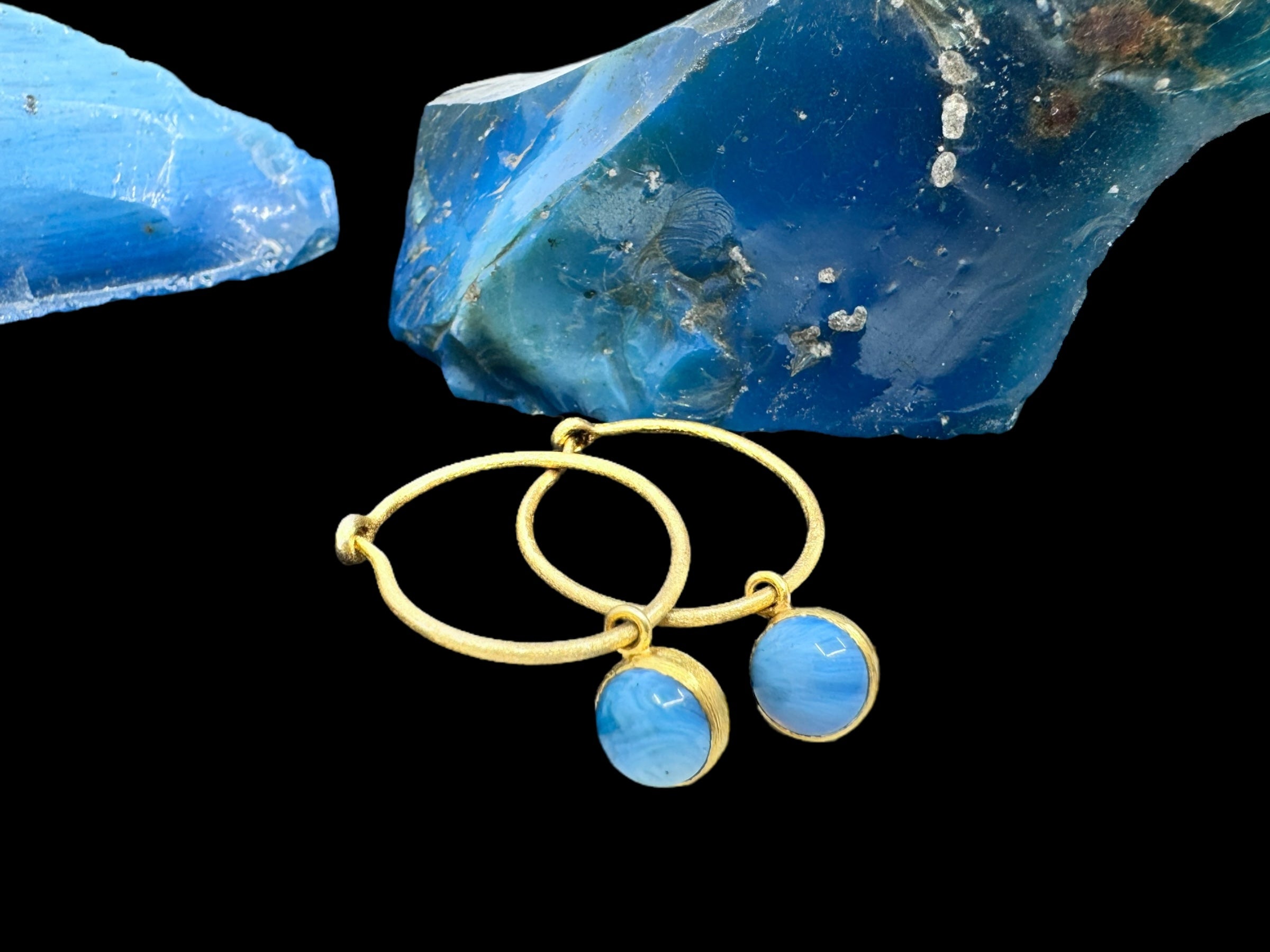 Cirkel Gold - Swedish Blue earrings featuring gold-plated hoops with blue gemstone pendants are elegantly displayed in front of two large Swedish Blue stones on a black background.