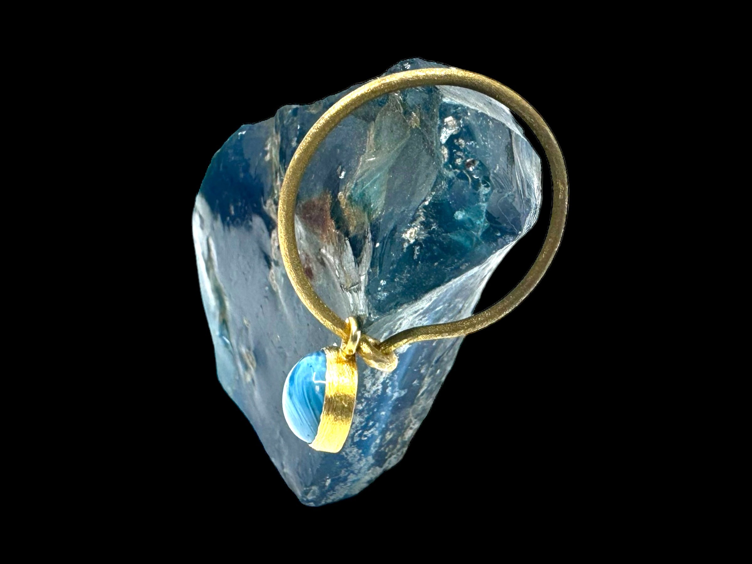 A gold-plated earring from the Cirkel Gold collection, featuring a Swedish Blue stone, is placed against a larger, dark blue crystal-like object set against a black background.