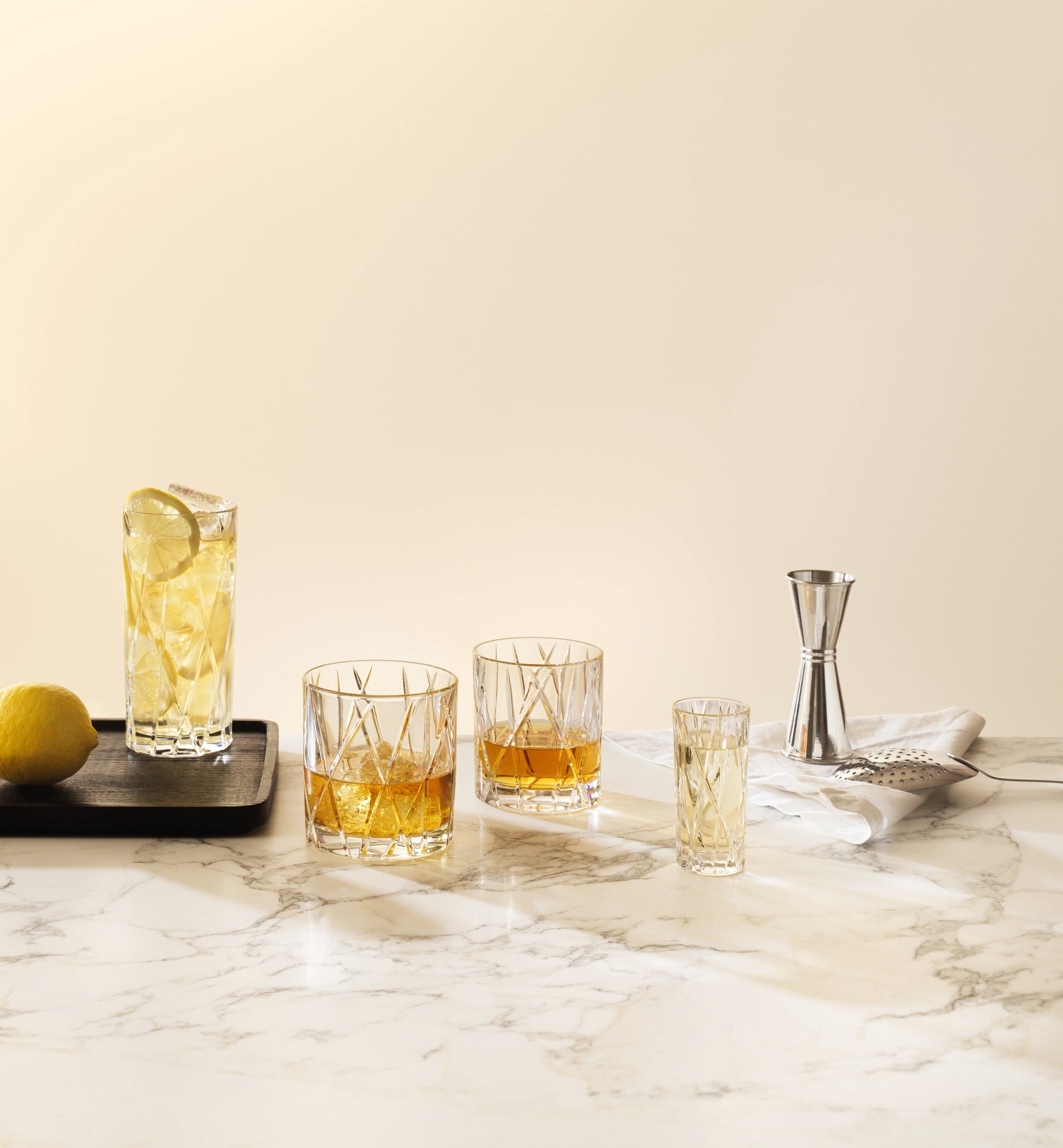 A highball glass with a lime wedge, two Orrefors City Double Old Fashioned Glasses filled with amber liquid, a shot glass, a lemon on a tray, and a jigger rest elegantly on the marble surface.