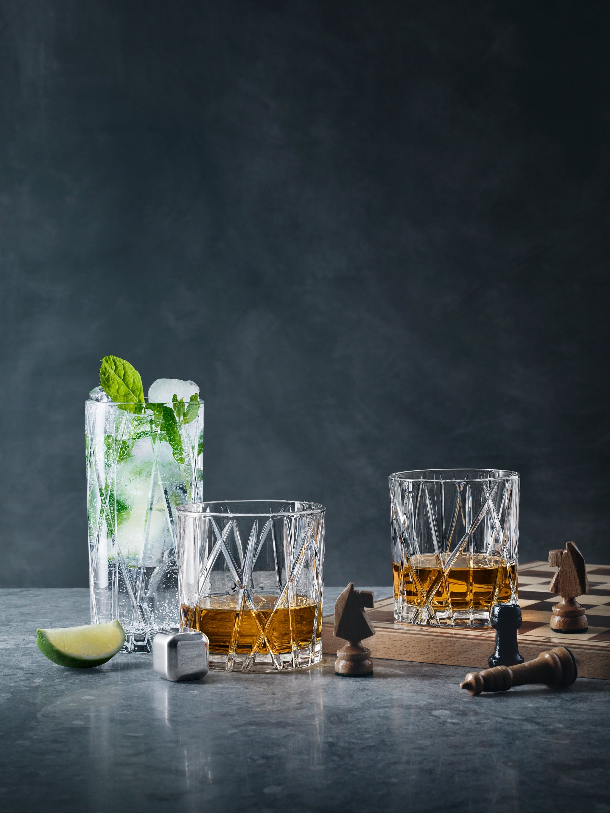 Two whiskey glasses, including the elegant Orrefors City Double Old Fashioned Glass from a 4-pack set, sit beside a mojito and chess pieces on a gray surface.