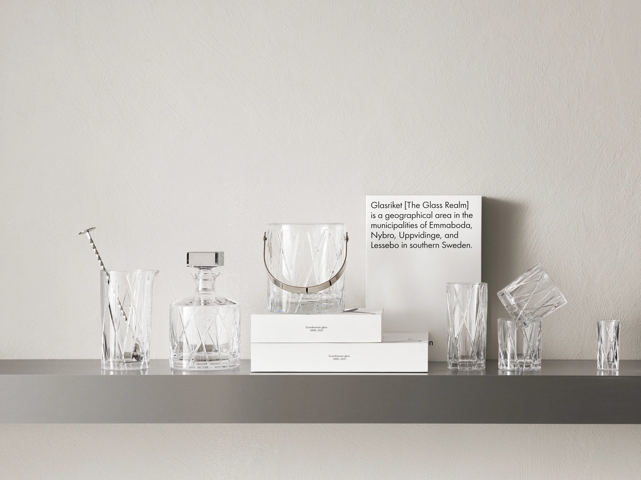 A stunning collection of crystal glassware graces the shelf, featuring Orrefors pieces like a decanter and City Double Old Fashioned Glasses (11oz, 4-Pack) by Martti Rytkönen, reflecting insights about Glasriket, a renowned region in southern Sweden famous for its exquisite glass artistry.