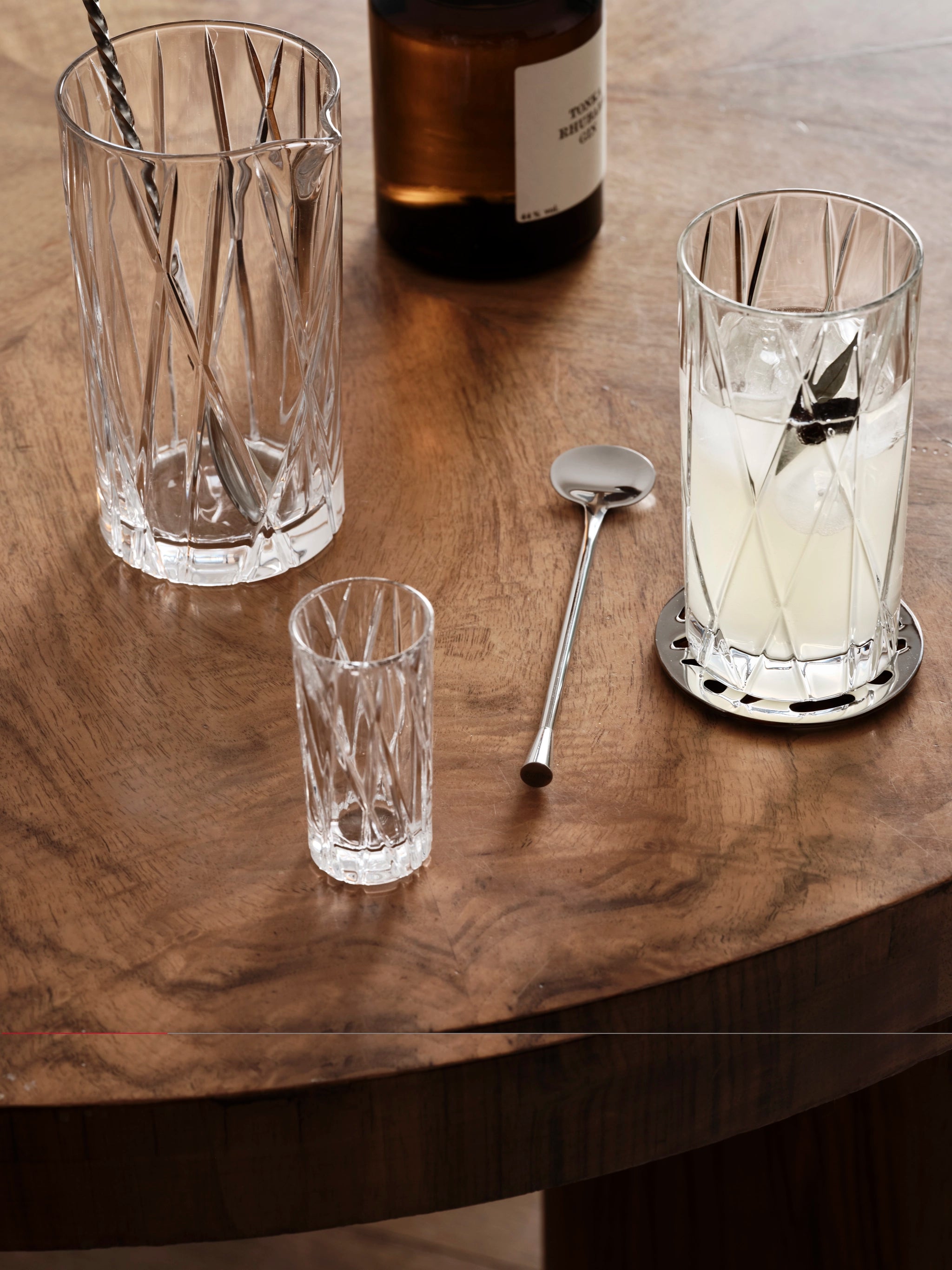 Orrefors City Shot Glasses adorn the wooden table, paired with a metal stirrer and an icy drink in a Crystal Glass. A brown bottle quietly completes the sophisticated scene.