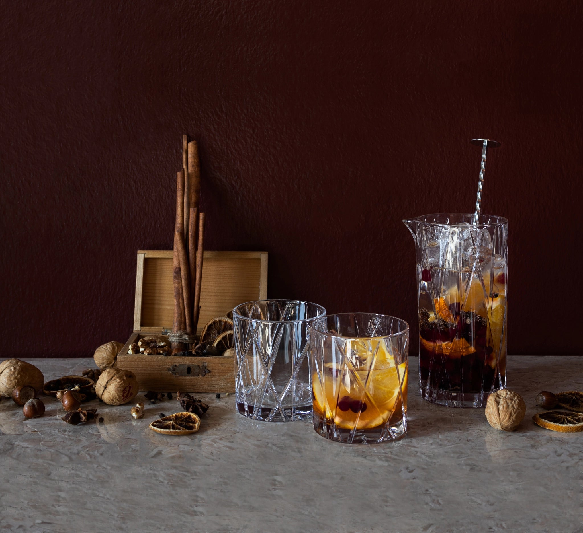 The Orrefors City Mixing Glass with Spoon, filled with fruit and liquid, is elegantly displayed alongside cinnamon sticks, nuts, and dried citrus on a marble surface.