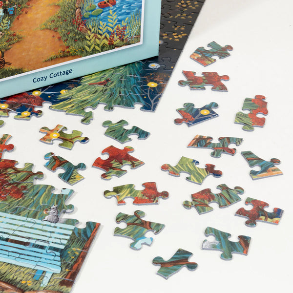 A partially completed PuzzleTwist Something's Amiss! Cozy Cottage 500 Piece puzzle by artist Mary Charles lies on a white surface, with several loose pieces spread nearby and its box visible.