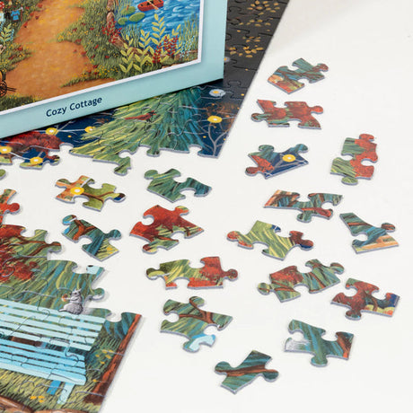 A partially completed PuzzleTwist Something's Amiss! Cozy Cottage 500 Piece puzzle by artist Mary Charles lies on a white surface, with several loose pieces spread nearby and its box visible.