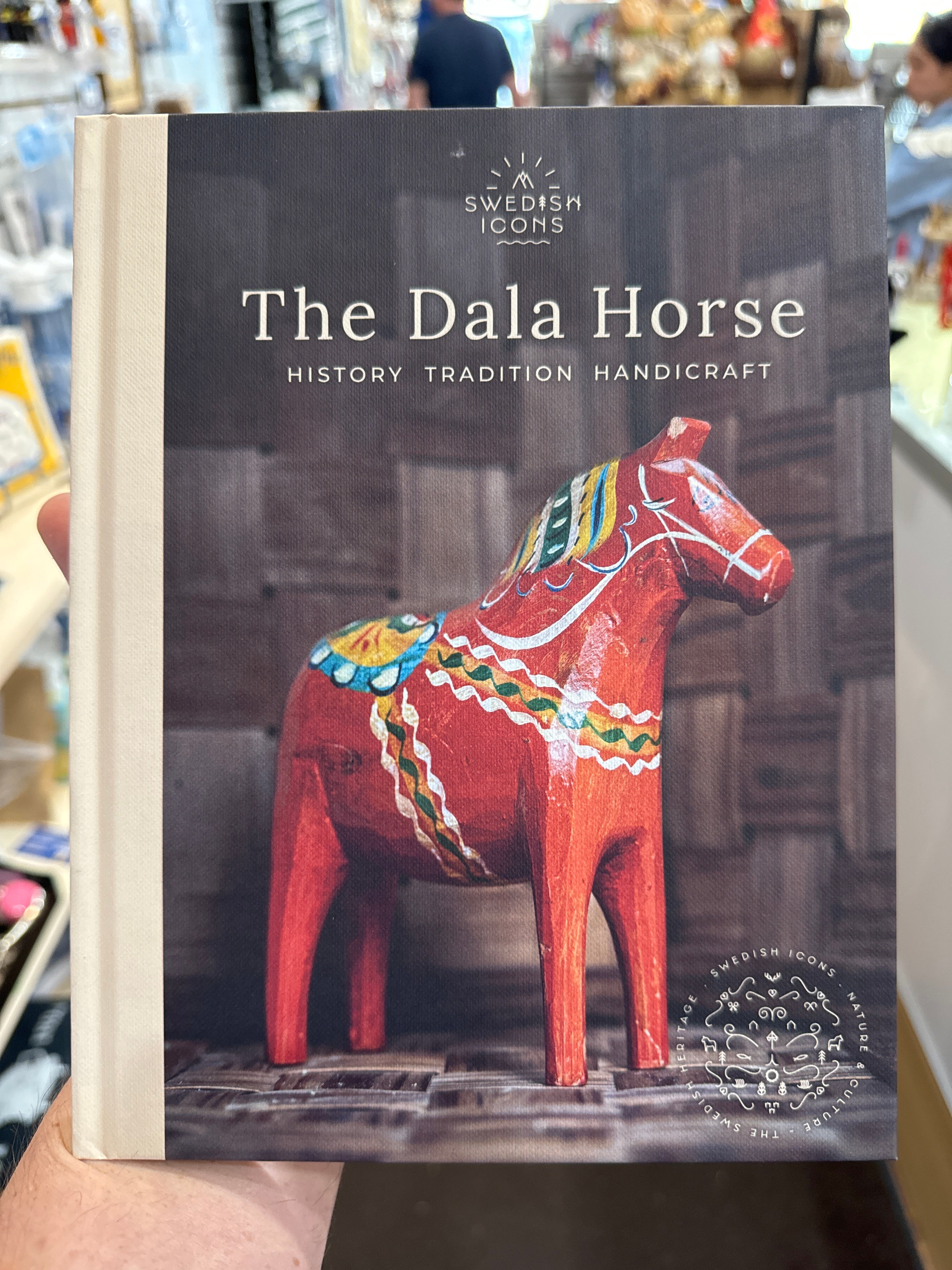 A person holds the book *The Dala Horse, History Tradition Handicraft*, which showcases an image of a traditional Swedish Dala Horse from Dalarna, intricately carved in vivid red with colorful patterns.