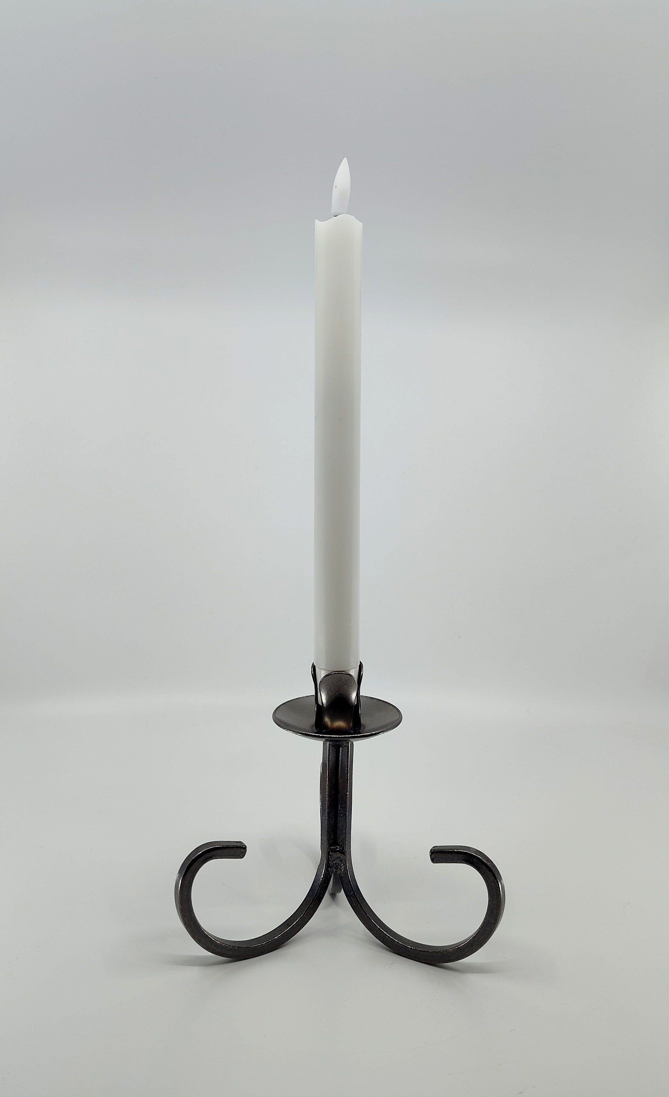 A tapered white candle elegantly stands in a Danish Iron Taper Candleholder with a Curly Bottom, set against a plain gray background.