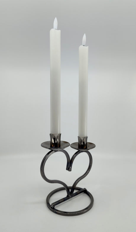 Two white candles elegantly sit in a hand-forged Danish Iron Taper Candleholder, featuring a large heart design for two taper candles, set against a plain background.