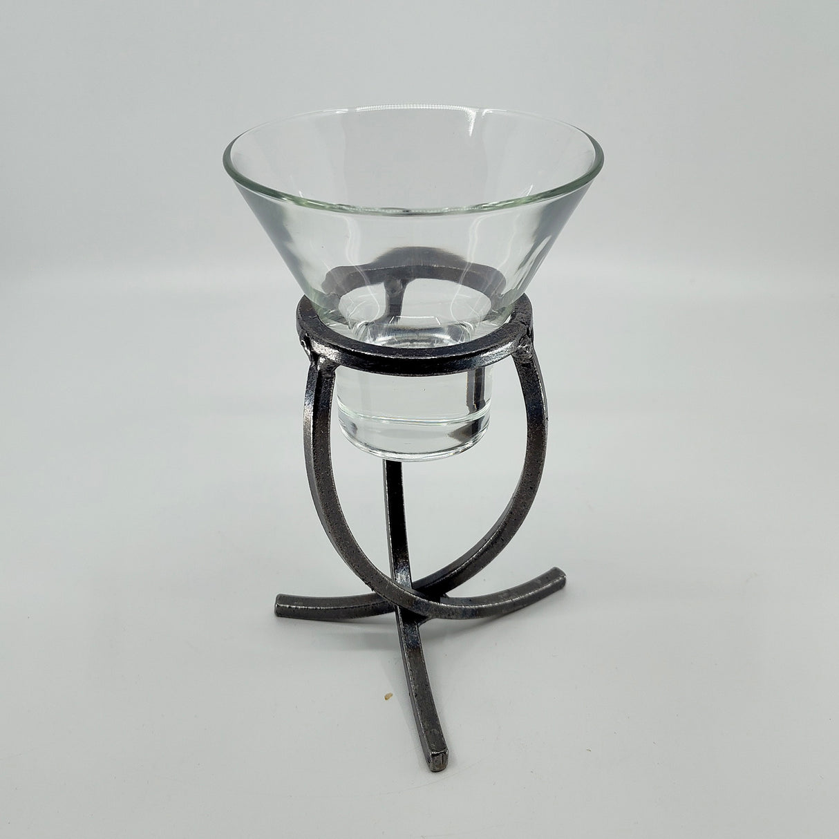 A clear glass bowl rests elegantly on a Danish Iron Candle Holder with a crossed iron base and cup for a ball candle, set against a plain white backdrop.