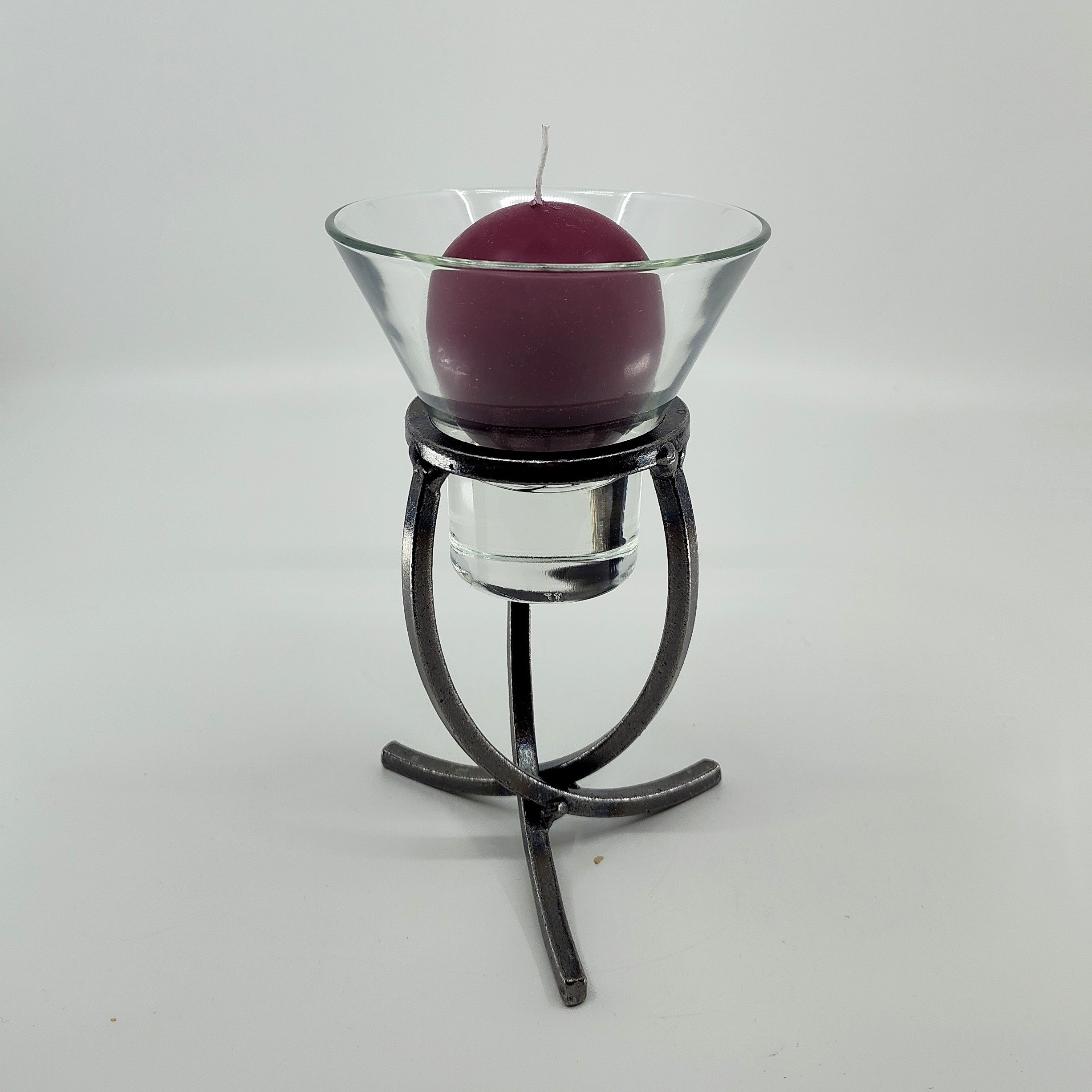 A purple candle housed in a Leif Cup glass sits on a Danish Iron stand with a crossed iron base and three legs, set against a white background.