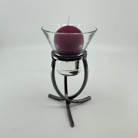 A purple candle housed in a Leif Cup glass sits on a Danish Iron stand with a crossed iron base and three legs, set against a white background.