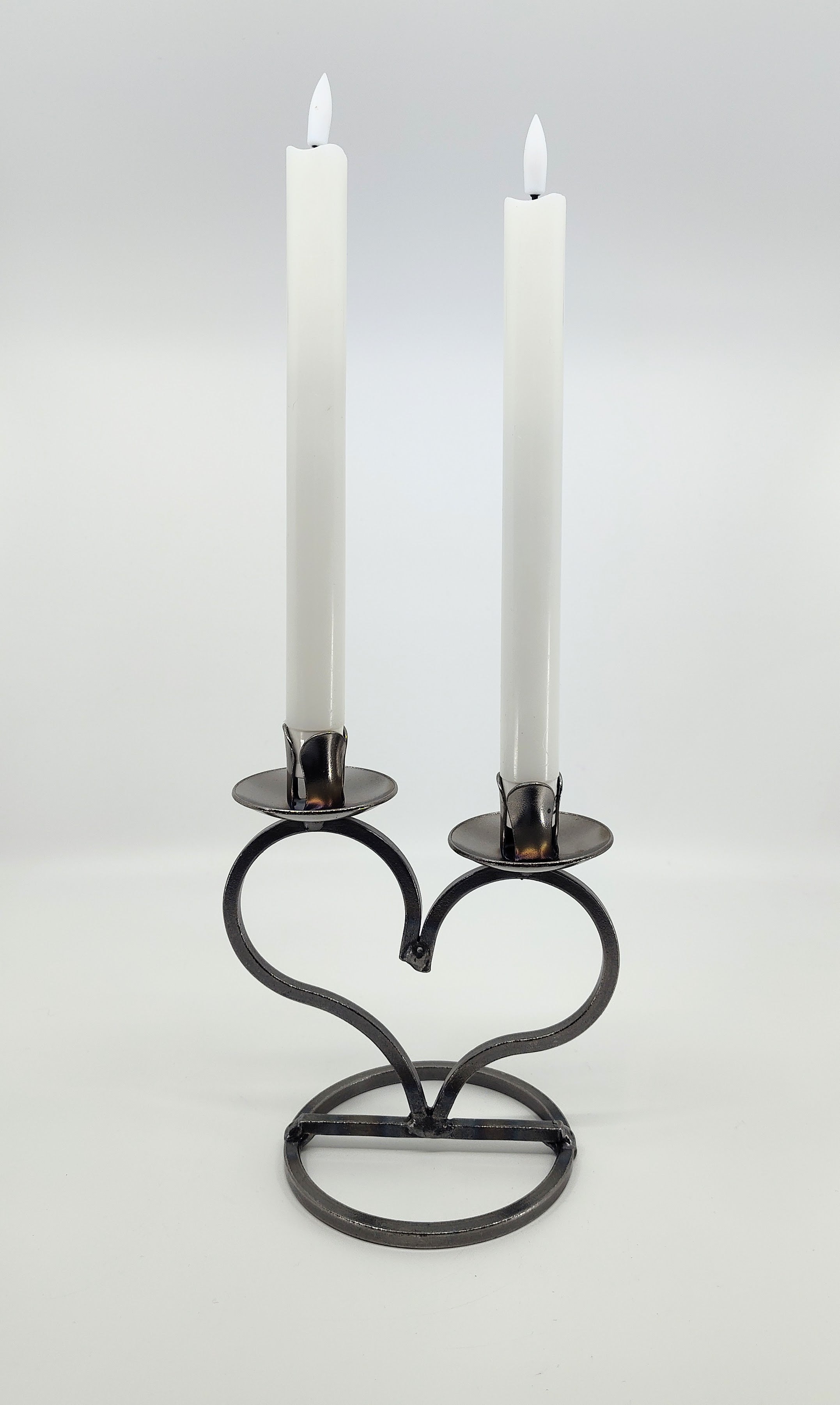 Two lit candles rest elegantly in a Danish Iron Taper Candleholder with a large heart, showcasing Scandinavian craftsmanship, against a plain white background.