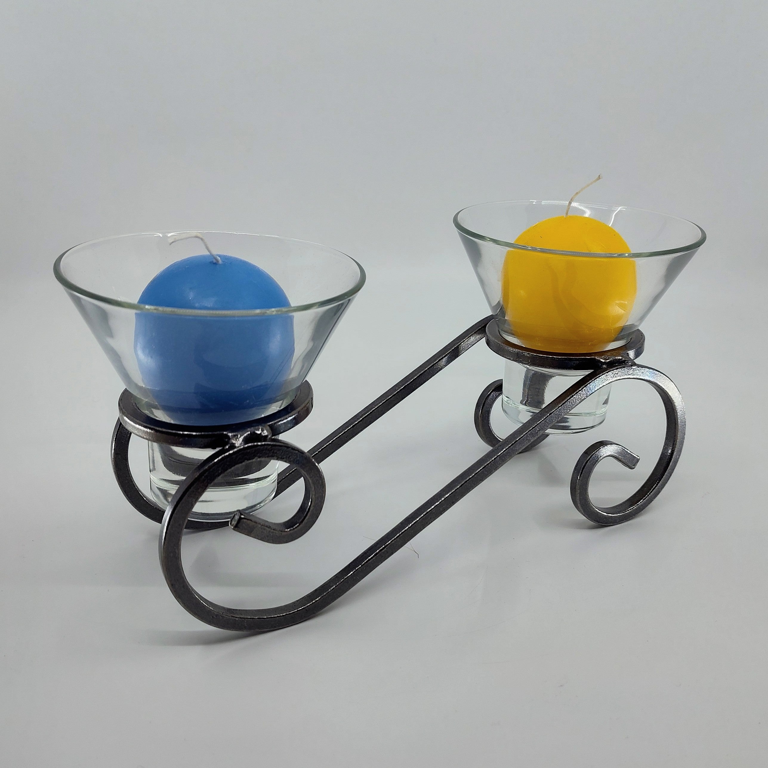 A Danish iron candleholder elegantly supports two Leif glass cups, each holding a blue and yellow candle against a crisp white backdrop.